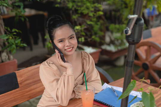 A smiling young lady is talking on the phone with a client on an al fresco restaurant while preparing for her upcoming meeting.