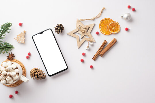 Top view photo of smartphone wood star ornaments cup of cocoa with marshmallow pine cones spruce branch mistletoe berries cinnamon and dried citrus slices on isolated white background with copyspace