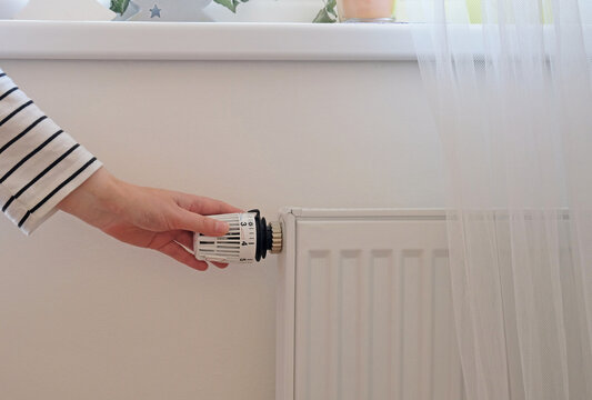 Woman regulates temperature at home with the heating thermostat to save energy, close-up with hand. Temperature control valve on radiator. White heater on wall with curtain in modern apartment.
