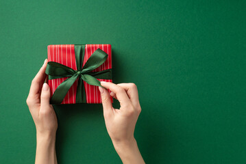 Christmas Day concept. First person top view photo of young woman's hands untying ribbon bow on red giftbox on isolated green background with empty space - obrazy, fototapety, plakaty