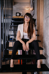 Obraz na płótnie Canvas Young Italian brunette with loose hair dressed elegantly sitting on the steps in office, needs a brake. Vertical shot of beautiful caucasian girl in glasses exhausted after work. Lawyer at office
