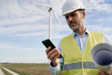 Caucasian male engineer standing on wind turbine field and using mobile phone