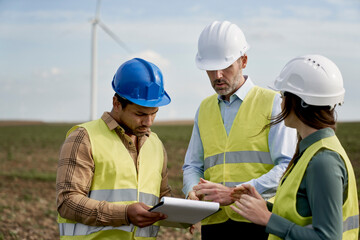 Team of caucasian and latin engineers standing on wind turbine field and discussing over documents