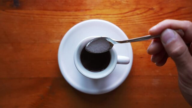 Down shot of a hand that is putting suger in a hot espress cup and stirs, on a brown wooden Table. Steam is rising up.