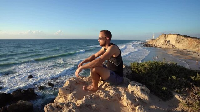 A man in sports wear and black sunglass sitting in rocky beach and looking at the blue ocean in Haderah, Israel.