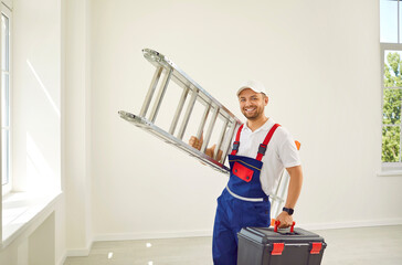 Portrait of a happy male worker in the house. Man in a work uniform jumpsuit standing in an empty white room at home, holding a ladder and a toolbox, looking at the camera and smiling. Repairs concept