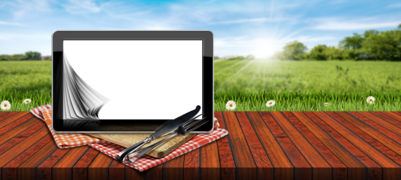Tablet computer with blank pages on a wooden cutting board, red and white checkered tablecloth, silver cutlery. On a wooden table against a blurred countryside landscape with green grass and flowers.