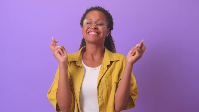young superstitious curly African American woman in hope of fulfilling wish clenches fingers with cross and closes eyes waiting for results of interview or exams, stands in purple studio