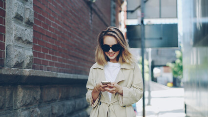Pretty blond girl is using modern smartphone touching screen walking in city and smiling enjoying device. Young woman is wearing fashionable clothing and trendy sun glasses.
