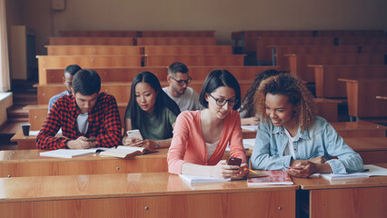 Young people are using smartphones and talking sitting at desks in lecture hall at university....