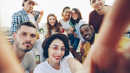 Obraz premium Young woman is taking selfie holding camera and looking at camera while her friends are clinking glasses, posing and showing hand gestures at funny outdoors party.