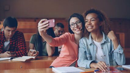 Cheerful girls students are taking selfie with smartphone sitting at tables at college, young women are posing making hand gestures and hugging. Friends and technology concept.