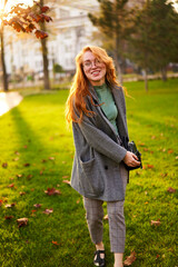 Redhead smiling pretty woman walking in park on sunny autumn day. Elegant female poses in stylish authentic outfit has fun laughing on green lawn with vivid foliage, leaves. Girl enjoys fall season.