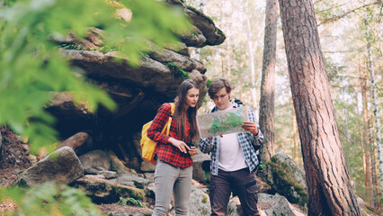 Lost tourists are standing in forest near huge rocks, looking at map and talking discussing way to their destination place. People, hiking and nature concept.