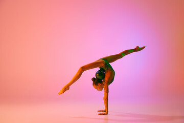 Fototapeta premium Young flexible teen girl rhythmic gymnast in motion, action isolated over pink background in neon light. Sport, beauty, competition, flexibility, active lifestyle