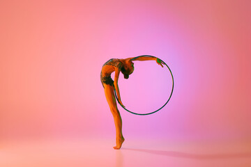 Young flexible teen girl rhythmic gymnast in motion, action isolated over pink background in neon...