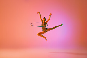 Young flexible teen girl rhythmic gymnast jumping isolated over pink background in neon light. Sport, beauty, competition, flexibility, active lifestyle