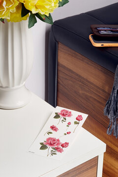 Close-up shot of a sheet of 6 beautiful colored temporary rose tattoos lying on a nightstand with a vase of flowers. The sheet of water-based temporary tattoos is in the room. Top view.
