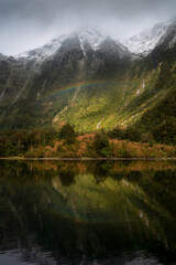 Naklejka na ściany i meble Sunlight breaks through the clouds in the Doubtful Sound revealing a rainbow up against the snowcapped peaks of the fjord.