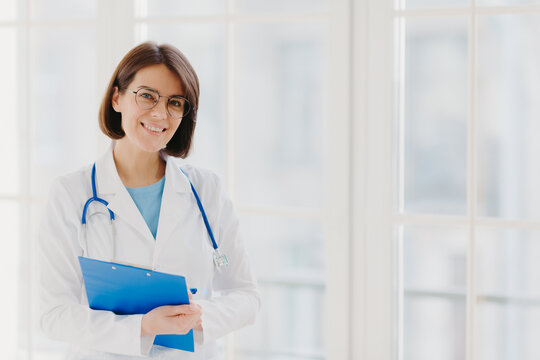 Good looking female expert creats medical prescription, writes down information in clipboard, thinks about good advice for patient, stands indoor, copy space for your information. Medicine concept