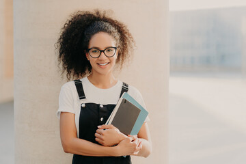 Adorable curly haired female student wears white casual t shirt and overalls, holds notepad or textbook, looks through transparent spectacles, stands against white background. People and lifestyle