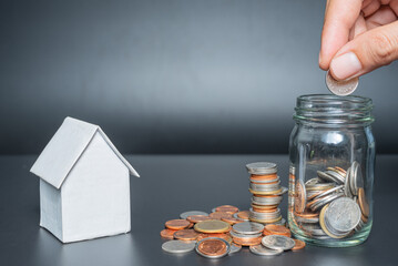 Hand putting Thailand coins in jar with heap coins on black background with house model