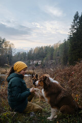 Obraz premium Owner holds paw of Australian Shepherd and smiles. Spend time with animals in nature. German Shepherd sitting behind. Young Caucasian woman sitting with dog in autumn forest by river. Hiking with dog.