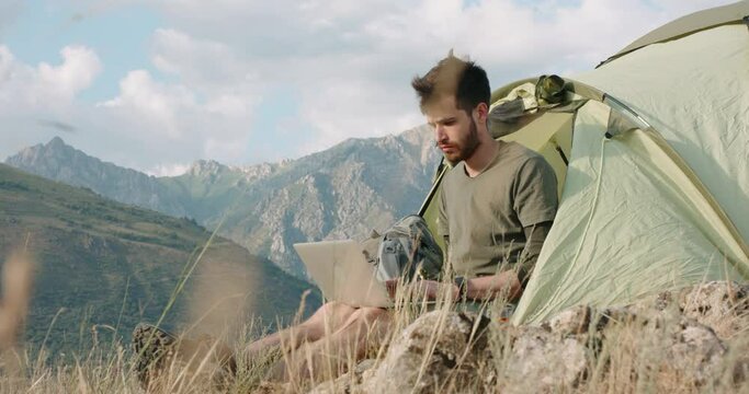 A modern millennial young man is sitting near a tent in a mountainous area, working on a laptop remote from the office. The copywriter writes the text for the site. The new lifestyle of Generation Z