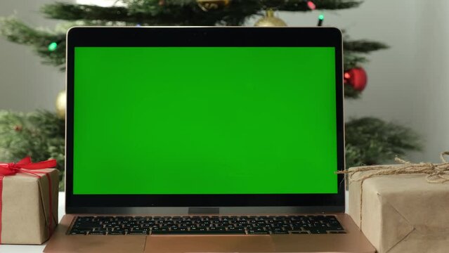 Close up modern laptop with green screen surrounded by gifts on background of Christmas tree. Online gift shopping for Christmas concept. Chroma key computer on the table. New Year holidays