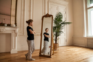 Alone cute teenager girl at mirror in living room at home, looking her. Sad lonely child stands and...