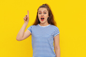 Excited young woman with brown-haired pointing finger up, looking at camera with open mouth, have a cool idea, standing in white-blue striped t shirt over yellow background