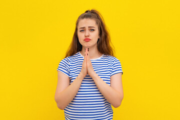 Calm beautiful young woman keeps palms pressed together, showing pray, namaste gesture, express gratitude, thank you sign, standing in white-blue striped t shirt over yellow background