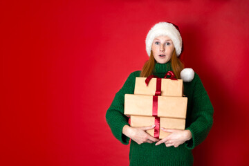 Merry Christmas Portrait of a beautiful young teenage girl in a cozy knitted green sweater and Santa's hat holding gift boxes. The red background is the place for the text.