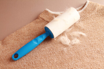Cloth fabrics cleaner lint roller brush with paper roll blue grip on brown beige color sweater background. sticky roller, pet cat dog fur dust hair remover. Close-up