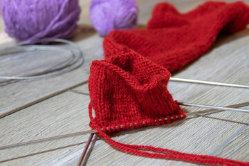 Sock in the process of knitting with needles and yarn of red color on a gray wooden table