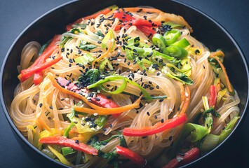 Japchae or  Chapchae in black bowl on the dark table. Korean cuisine glass chapchae noodles dish with vegetables and sesame. Asian traditional food. Authentic meal, close up