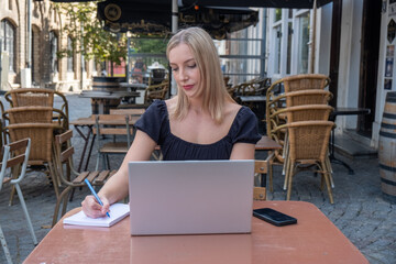 Attractive young blonde business woman using her laptop while sitting outdoors in a European city at the typical Belgian cafe terrace. High quality photo