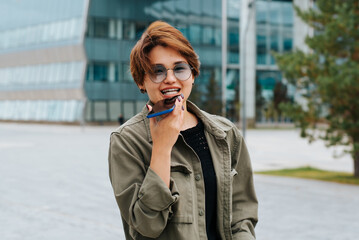 Cheerful laughing Asian woman is recording voice message on a smartphone outdoors. Portrait of...