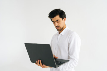 Studio shot of serious young man in shirt standing with laptop and typing on keyboard, doing freelance job, writing email on white isolated background. Focused male posing with pc looking at screen.
