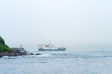 fishing vessel emerges from behind a cape with a lighthouse, sailing into a foggy sea