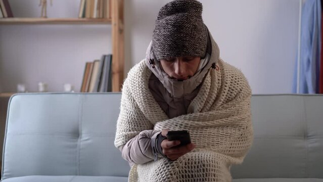 Sad young man sit at the table at freezing cooled flat in warm cap and blanket shiver tremble with cold, using smart phone. Unhappy guy spend time at home feel bad suffer of heating problems.