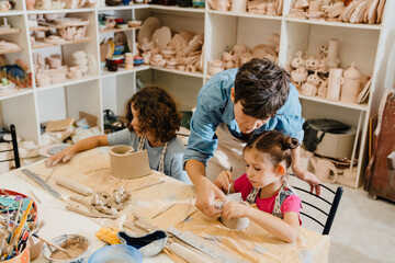 Mature female teacher sculpting with kids and assisting them at ceramics workshop