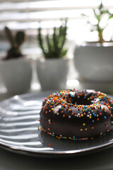 a sweet chocolate donut with multi-colored sprinkles on top lies on a gray plate. in the background are homemade flowers in white pots