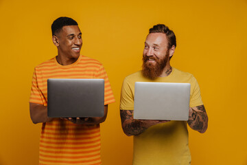 Two excited men holding laptops and smiling at each other while standing isolated