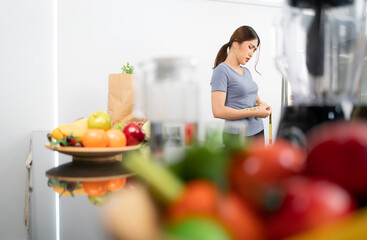 Asian woman measuring her waist with worry and nervous emotion with blur fruits and vegetables in the foreground. Wearing sporty clothes. Image with copy space.