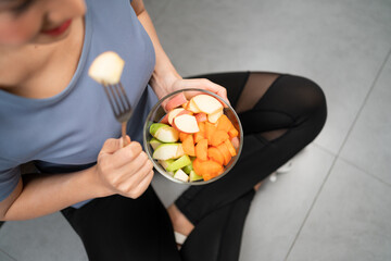 Top view of active woman sitting and eating cut red and green apple mixed with carrot in glass bowl picking with a fork. A dieting vegetarian meal.
