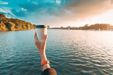 Takeaway cup with hot coffee in female hand against scenic sunset lake background