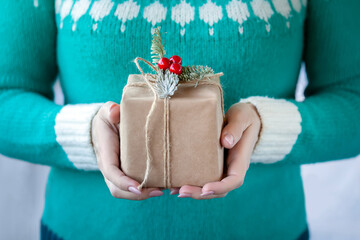 A woman in a warm green sweater holds a paper box in her hands with a Christmas decor made from a fir branch and red berries. Close-up of female hands holding a Christmas or New Year's gift.