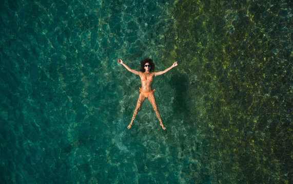 Aerial view of a beautiful woman wearing an orange bikini while floating and sunbathing in a transparent sea in San Sostene, Calabria, Italy.