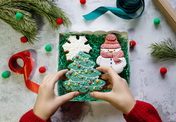 Girl's hands in red knitted sweater put gingerbread in the form of snowman into gift box with Christmas tree and snowflake gingerbreads next to pine branch, ribbons and wrapping paper. Christmas vibes